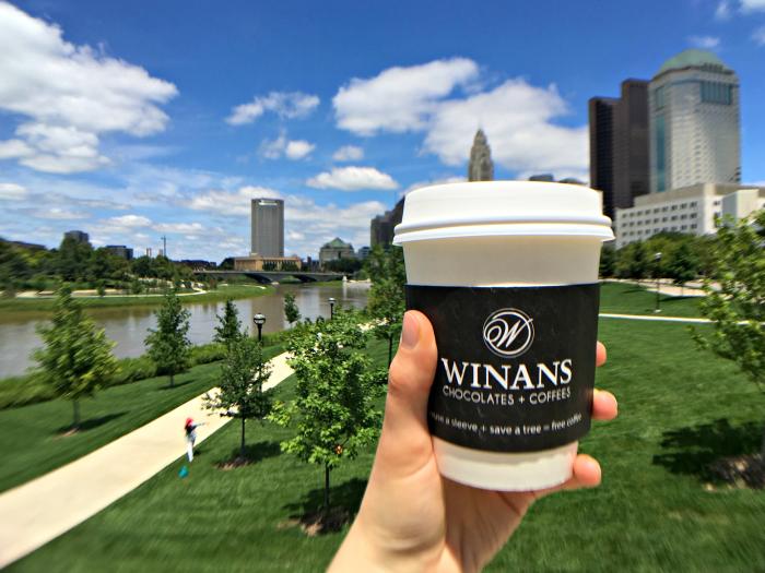 Hand holing to-go mug of coffee with Winan's label in front of Scioto Mile park