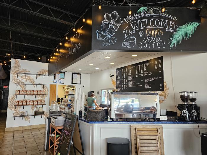 Interior of Angel's Island Coffee featuring a bright coffee counter, espresso equipment, menu boards, coffee bean grinders, and shelves stocked with bags of coffee. A large hand-drawn chalk mural reading "Welcome to Angel's Island Coffee" with tropical flowers and palm leaves decorates the space, while string lights hang from the open ceiling, creating a warm and inviting atmosphere.