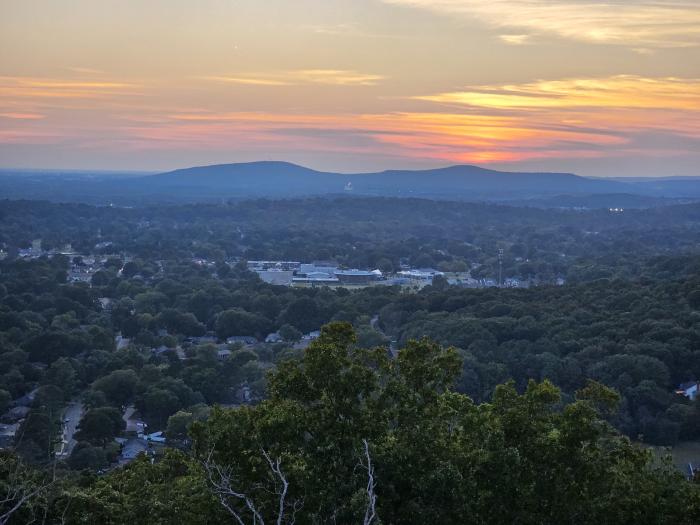 Panoramic sunset view from Blevins Gap overlooking Huntsville and the surrounding valley. Layers of tree-covered hills stretch into the distance beneath a colorful sky painted with shades of orange, pink, and blue as the sun sets behind the mountains.