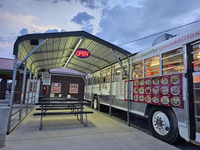 Evening view of the Taqueria El Cazador bus restaurant with illuminated menu boards, a glowing "Open" sign, and covered outdoor picnic table seating. Dramatic clouds fill the sky above the iconic converted bus, creating a welcoming atmosphere for outdoor dining.