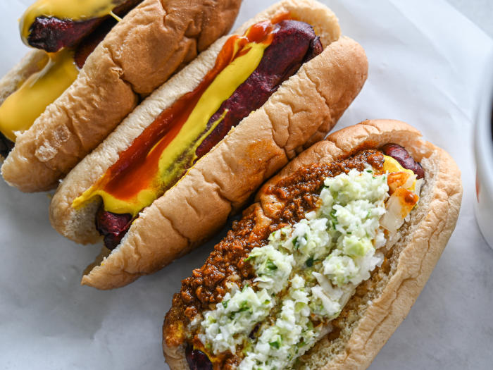 Three hot dogs with variety of ingredients including cheese, slaw, chili, and onions sit on a table.