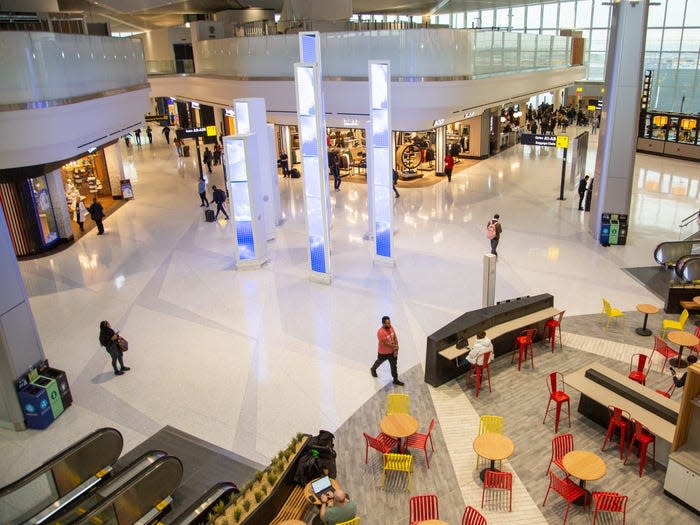 Overhead view of the spacious, modern interior of LaGuardia Airport Terminal B, featuring gleaming white terrazzo floors, illuminated rectangular blue columns, colorful red and yellow dining chairs, a food court seating area, retail shops along the perimeter, and travelers moving through the concourse.