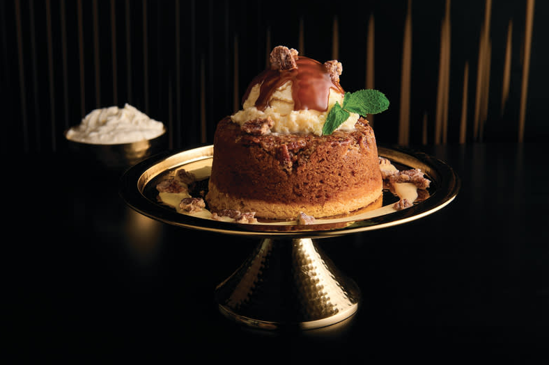 A personal-sized cinnamon-pecan butter cake rests on a antiqued gold cake stand. The cake is topped with a large scoop of butter pecan ice cream, caramel sauce, and candied pecans.
