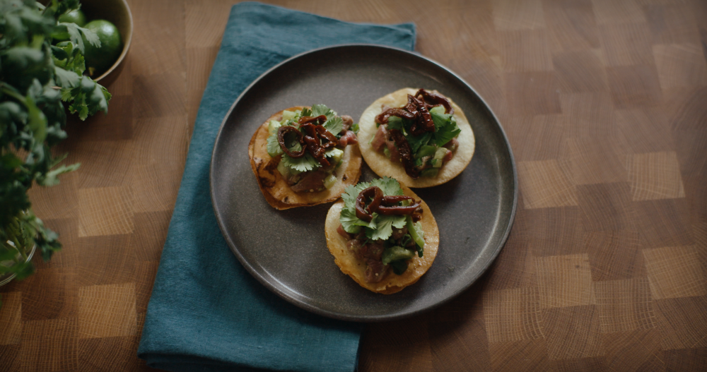 Three mini tostadas topped with thin slices of antelope ceviche, cilantro leaves and mini chipotle pepper rings on a dark grey stone plate.
