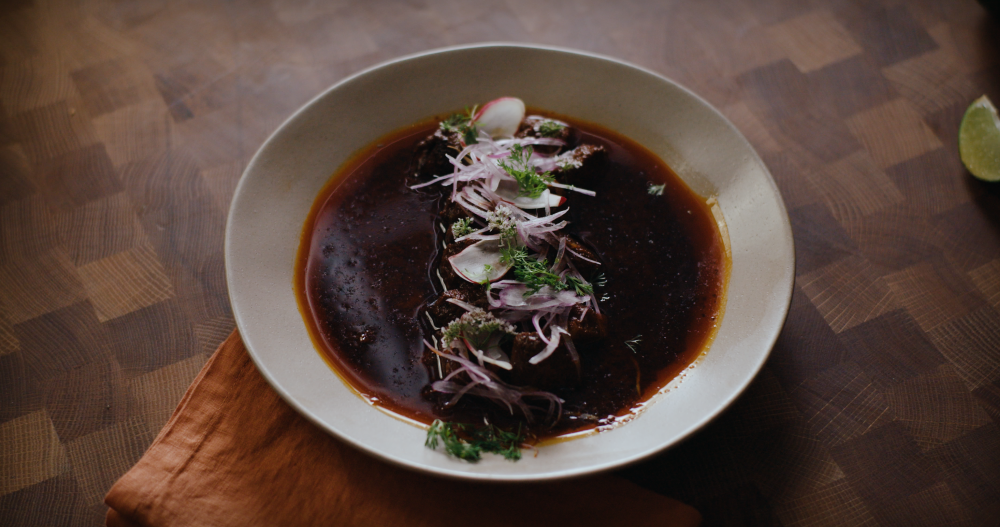 Line of stewed beef topped with delicate slices of radish, red onion and cilantro, surrounded by a rich-colored broth in a white bowl.