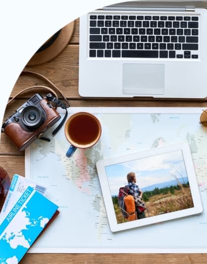 Top-down view of a wooden desk with a laptop, camera, coffee mug, airline ticket, and a tablet showing a person with a backpack overlooking a landscape, arranged on a world map to suggest travel planning.