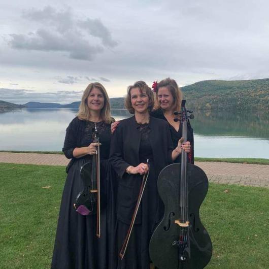 Three women playing string instruments dressed in black standing next to a lake