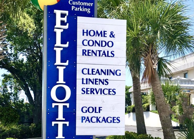 Elliott Beach Rentals' blue and white roadside sign in North Myrtle Beach, South Carolina, advertising home and condo rentals, cleaning and linen services, and golf packages, surrounded by palm trees under a clear blue sky.