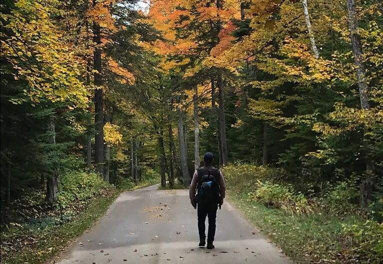 A man wearing a backpack walks along a paved road through the Mackinac Island forest of fall color