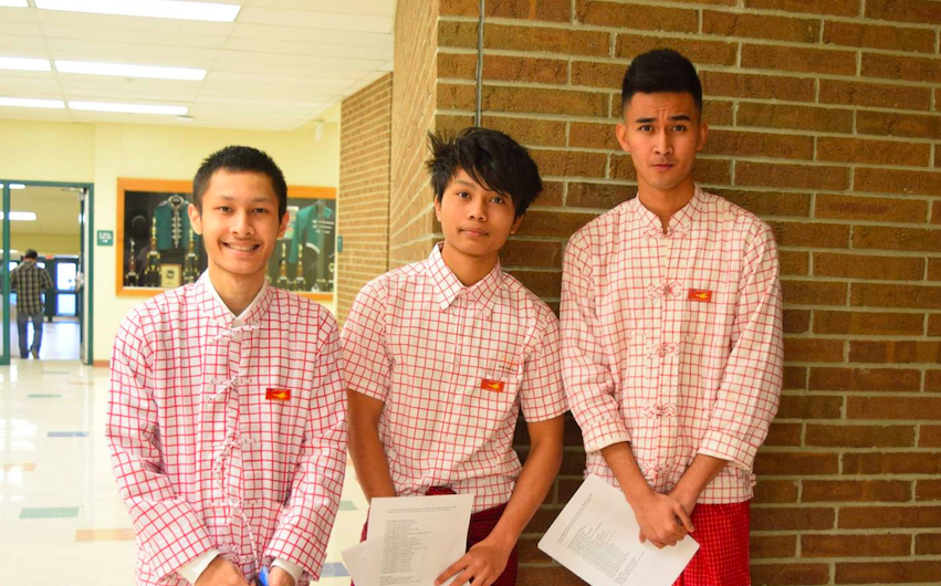 three boys standing together wearing white shirts with red criss cross pattern and burmese flag