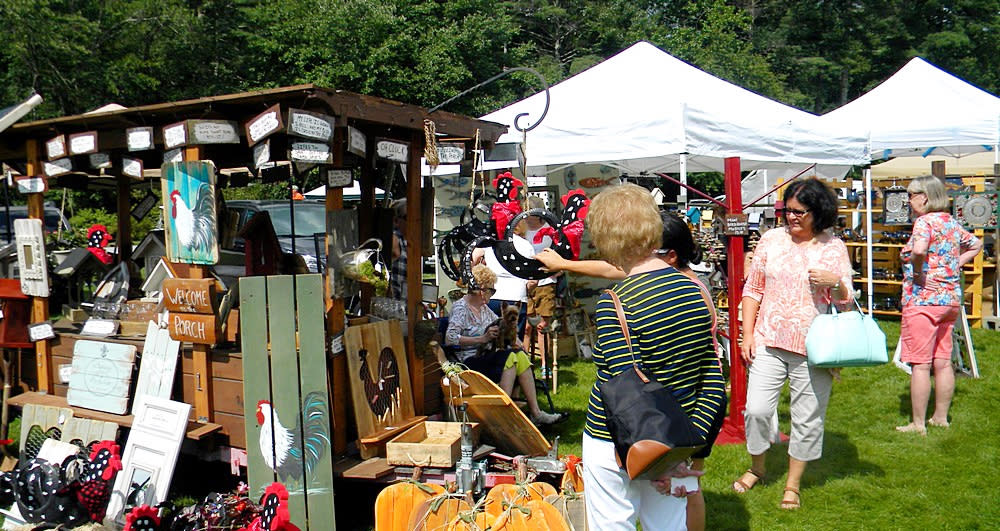 An outdoor craft fair takes place on a grassy lawn with multiple white vendor tents in the background. Shoppers, including a woman in a black and yellow striped shirt, browse rustic, country-themed items like painted rooster art and wooden pumpkins displayed on a custom wooden stand.