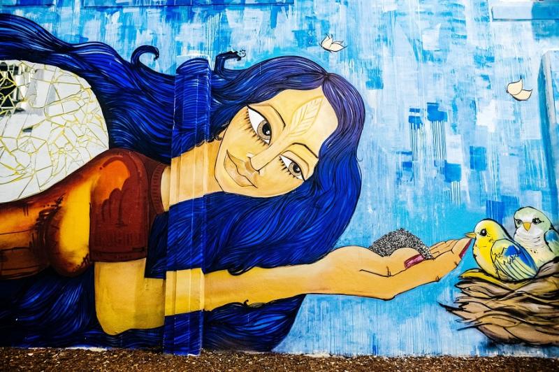Wall mural of a woman with blue hair, hand feeding a little yellow bird in a nest