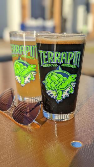 Two Terrapin beer glasses are shown, one filled with light beer and one filled with dark, sitting on a wooden table next to a pair of sunglasses.
