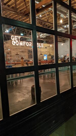 A light up sign that says "Get Comfortable" is viewed through the garage doors on the front of Creature Comforts Brewing Co.