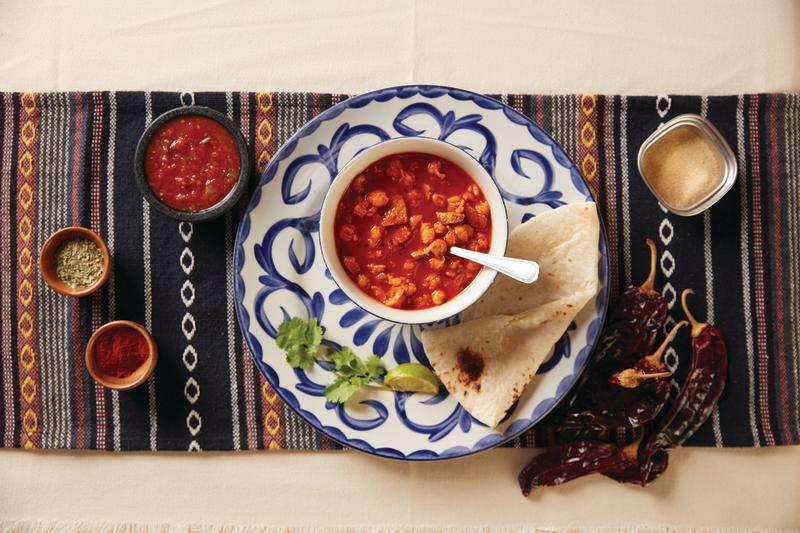 A bowl of posole sits on a blue and white plate with a tortilla on top of a New Mexican table runner adorned with New Mexican salsa and chiles