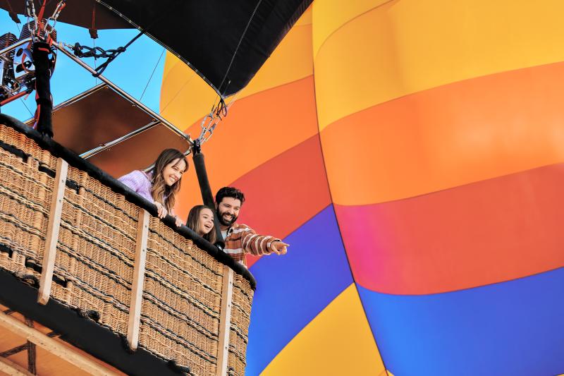 A man, woman and child ride in the basket of a hot air balloon. The man is pointing at something on the ground.