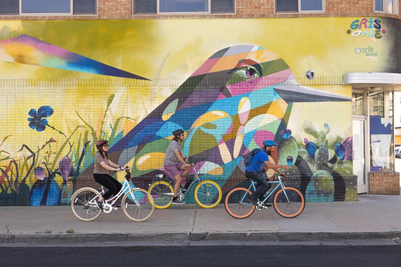 Three people ride by a mural on bikes in Albuquerque.