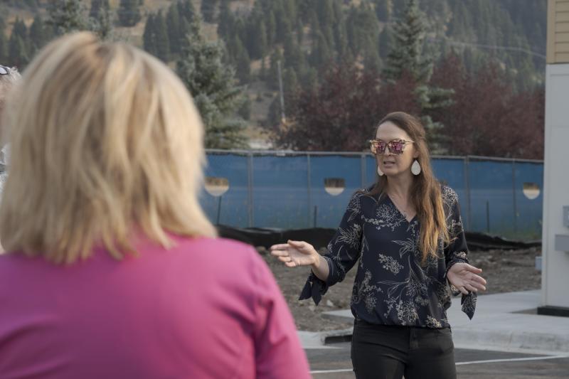 A woman in a patterned blouse gestures while speaking to another person, with a backdrop of trees and construction fencing.