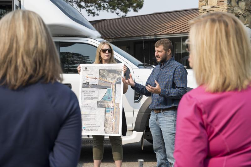 A man gestures while explaining a map held by a woman, surrounded by a group. A camper van and a rustic building are in the background.