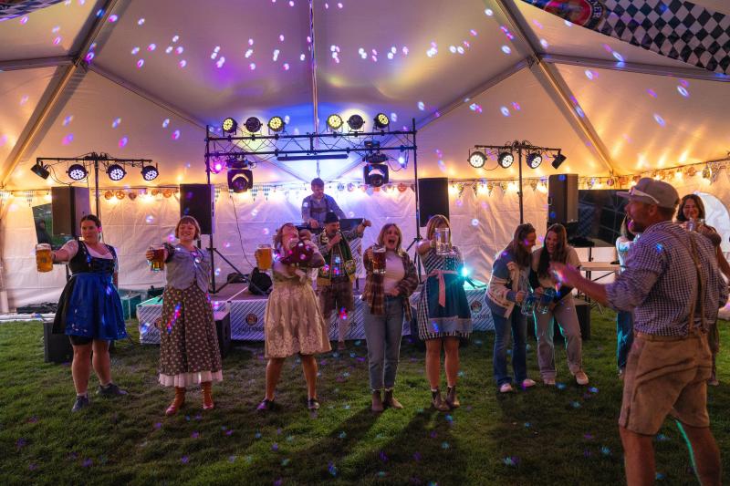 A lively outdoor event under a tent with colorful lights. Guests in festive Oktoberfest attire enjoy music, dancing, and drinks, creating a vibrant atmosphere.