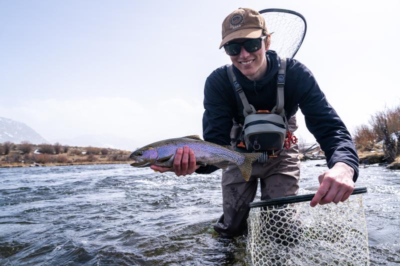 A young man in the river holding a trout with a fishing net in his other hand.