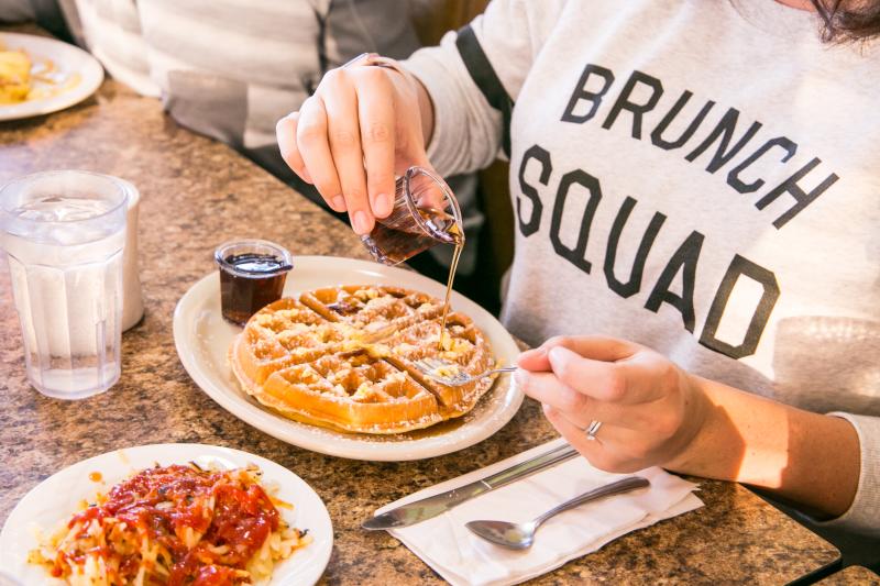 Person wearing a Brunch Squad shirt pouring maple syrup on a Belgian waffle at Cozy Table in Bloomington