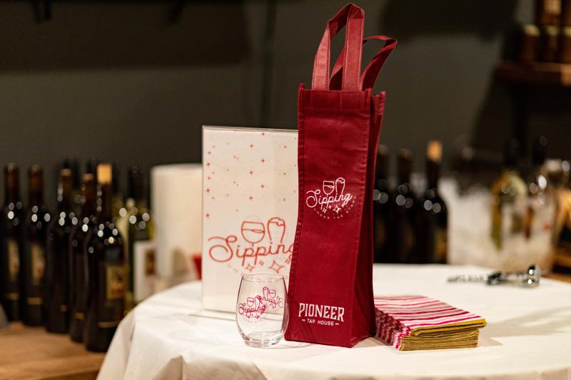 Photo of Sipping Under the Stars gear which includes a small wine glass and tote bag. Wine bottles can be seen in the background.