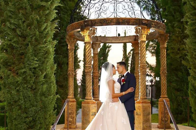 Bride in a ballgown and veil embracing groom in a blue tuxedo standing in front of a stone gazebo with Romanesque  pillars leading up to a round open top with metal swirls cut outs. Gazebo is surrounded on the sides by tall bushes that extend well above the gazebo's height.