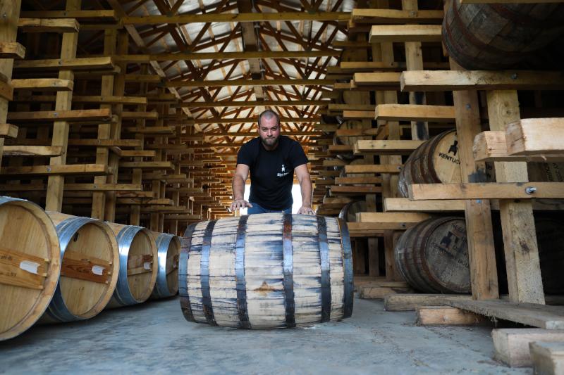 A man in a black t-shirt rolls a barrel towards the camera in the rickhouse of Neeley Family Distillery in Sparta.
