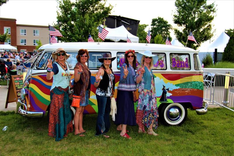 five women in hippie clothing giving peace sign stand in front of a brightly painted VW bus