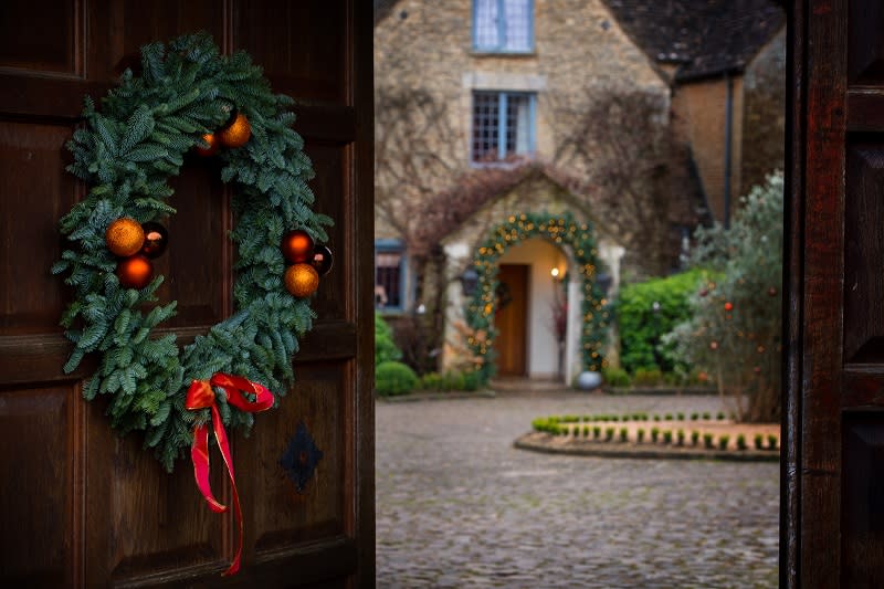 A Christmas wreath hangs on an ajar wooden door with a view of the front entrance and courtyard at Whatley Manor