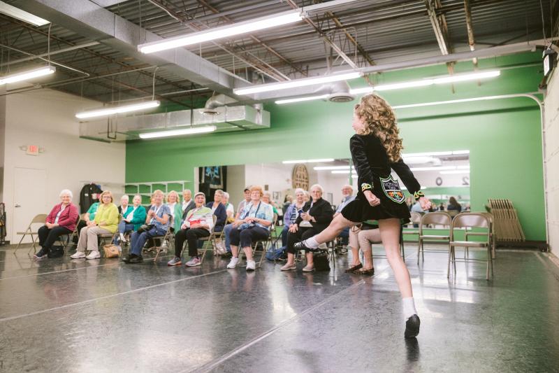Young Irish dancer performs in front of a seated audience in a studio with green walls and mirrors.