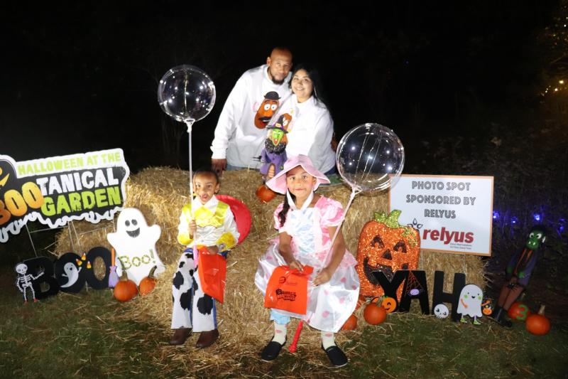 A family poses at a decorated Halloween photo spot during the Boo-tanical Garden event in Fayetteville. Two children in Toy Story costumes sit on hay bales holding clear light-up balloons and orange trick-or-treat bags, while two adults stand behind them smiling. The backdrop features pumpkins, ghosts, and a sign reading “Halloween at the Boo-tanical Garden.”