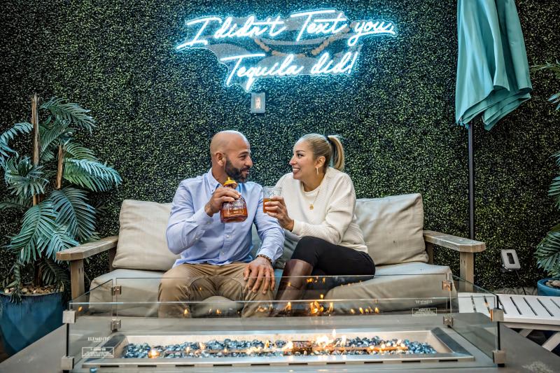 Couple toasting cocktails on a couch at a restaurant in front of a grass wall. Sign behind them on the grass wall says "I didn't text you tequila did"