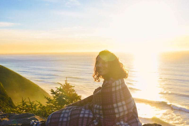 A woman sits on a cliff wrapped in a blanket near the coast at sunset.