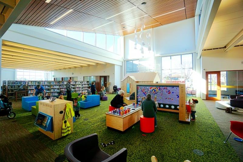 Children and adults in a the play area of the Brooklyn Park Library playing with a giant board with colored pegs
