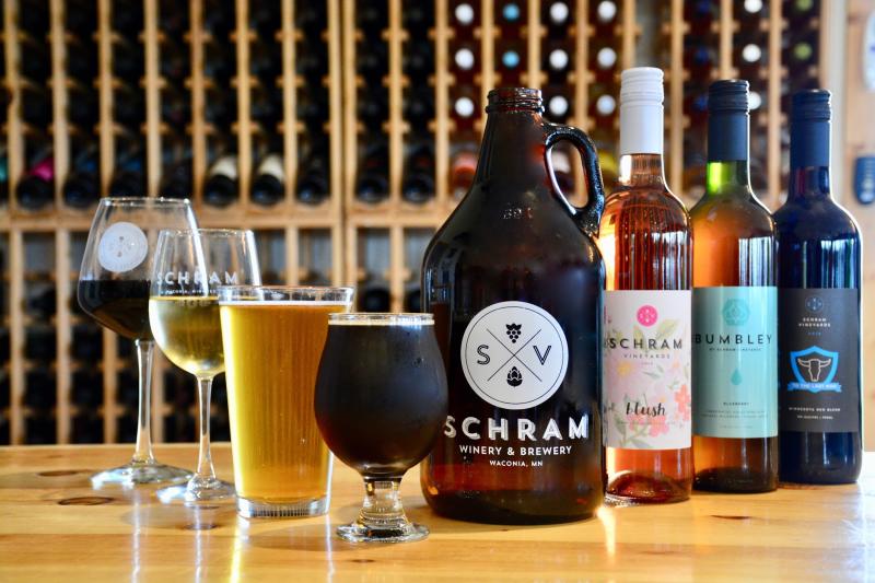 A variety of wine and beer glasses and bottles sit on a table in front of a large wine rack at Schram Vineyards Winery & Brewery