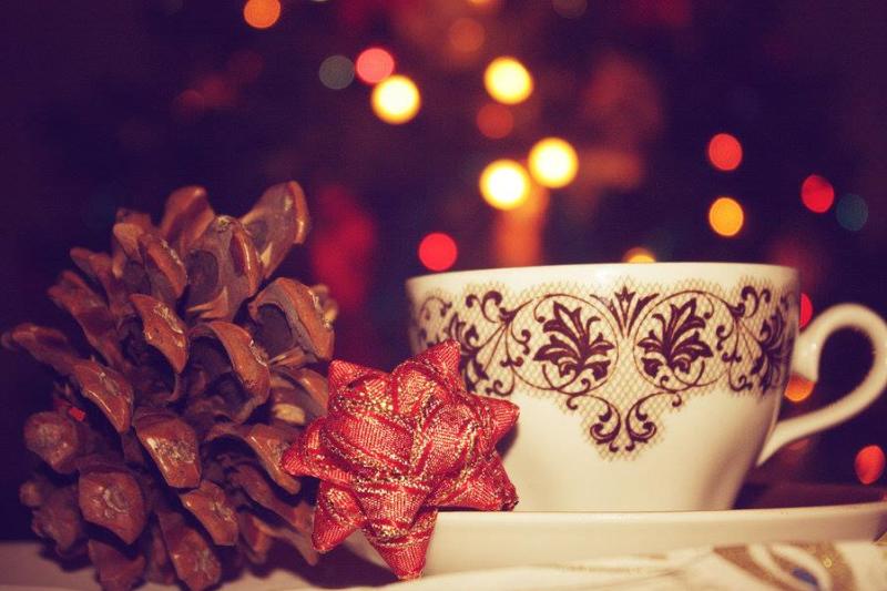 Tea cup sitting next to a pinecone and red ribbon with out-of-focus holiday lights in the background