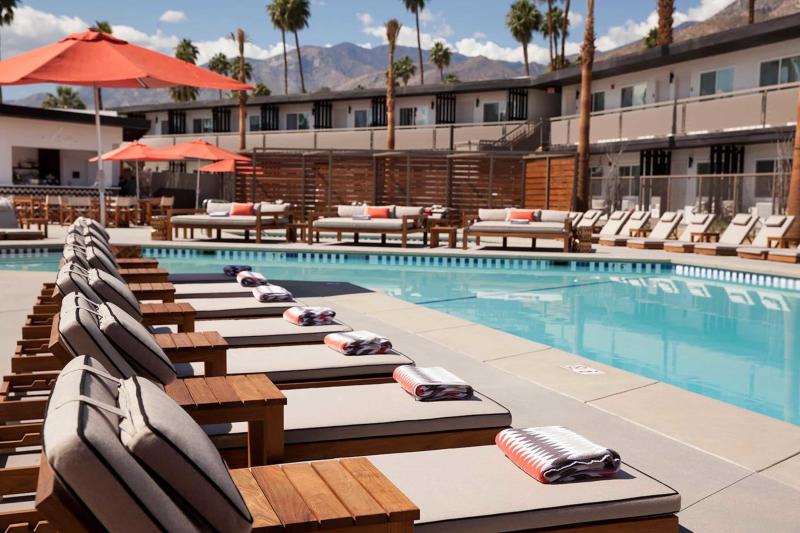 The V Palm Springs sunny poolside area with numerous sun loungers lined up next to a calm blue swimming pool, shaded by orange umbrellas, with palm trees and a mountain range in the background.