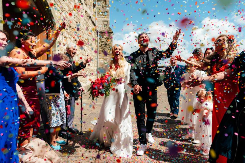 A bride and groom walking towards the camera with confetti all around them and guests either side