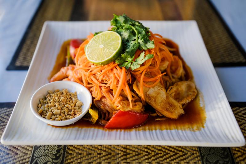 A plated Thai noodle dish with chicken, vegetables, shredded carrots, cilantro, lime, and crushed peanuts served on the side.