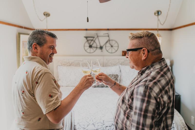 Two people smile and toast with glasses of white wine while standing in a cozy, well-lit bedroom at the Walnut Grove Bnb. The room features a neatly made bed with patterned bedding, warm lighting, and wall art of a bicycle above the headboard, creating a relaxed and welcoming atmosphere.