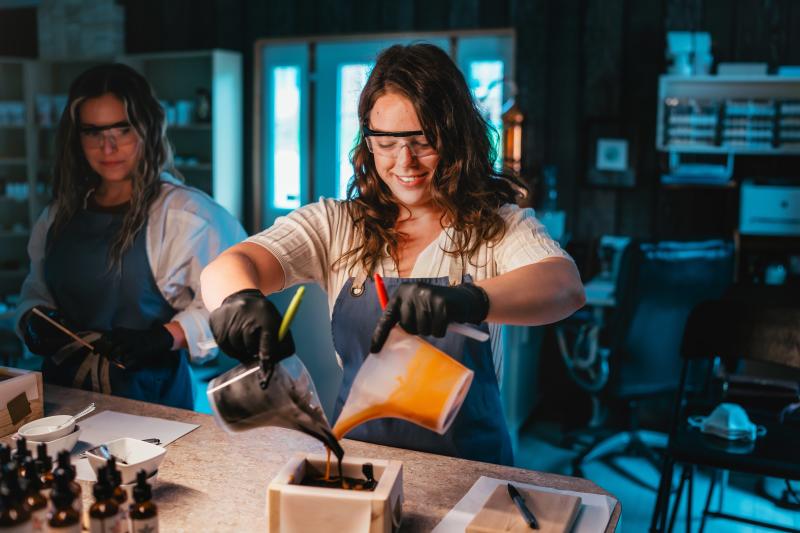 Two people wearing safety glasses and gloves pour orange and black liquid into a mold during a hands-on workshop.