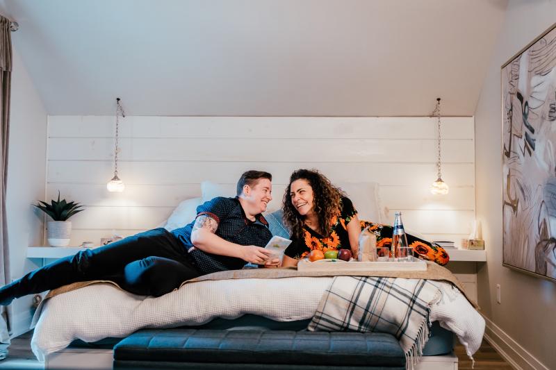 A couple relaxes on a neatly made bed in a cozy, modern bedroom at the Twisted Lemon Boutique Inn. They are smiling and looking at each other warmly while holding a book or pamphlet. A tray with fruit, water, and glasses rests on the bed in front of them. The room features soft lighting from hanging pendant lamps, a large abstract painting on the wall, and neutral decor with a blue bench and plaid blanket at the foot of the bed.