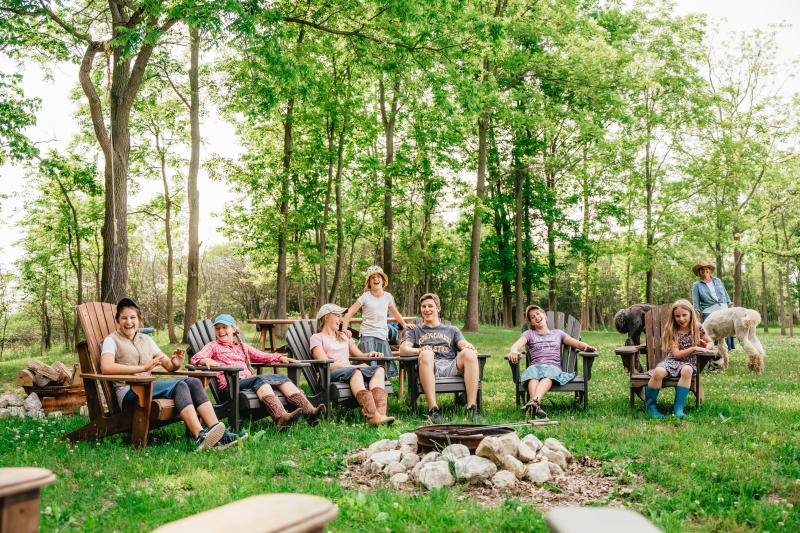 A group of people sit laughing in Adirondack chairs around a fire pit in a wooded outdoor space at Jireh Hills Family Homestead, with alpacas nearby.