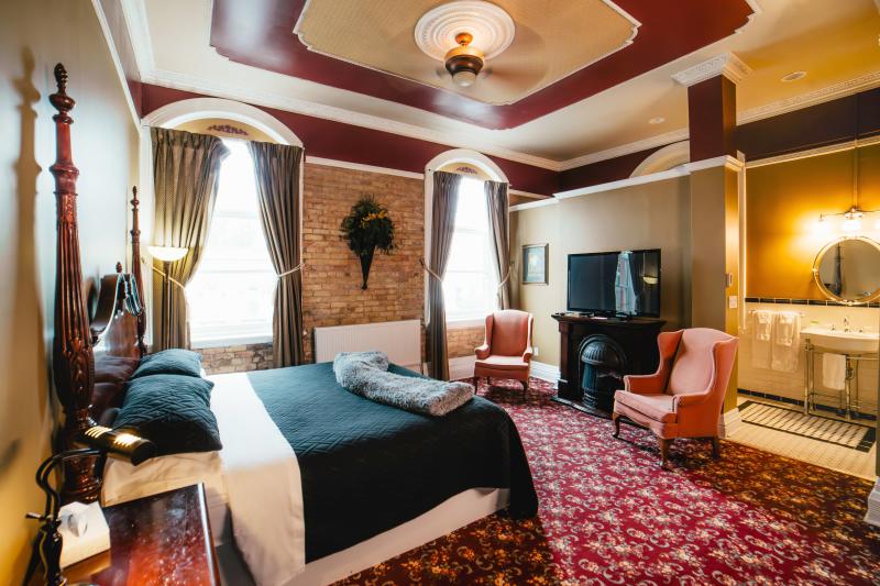 A warmly lit, elegant hotel room in the Clock Tower Inn featuring a large wooden four-poster bed with black bedding and decorative pillows. The room has tall arched windows with gold curtains, exposed brick walls, and red patterned carpet. Two pink armchairs sit near a fireplace and flat-screen TV, and a cozy bathroom with vintage-style fixtures is visible through an open doorway. The ceiling has ornate molding and a fan, adding to the room’s classic charm.