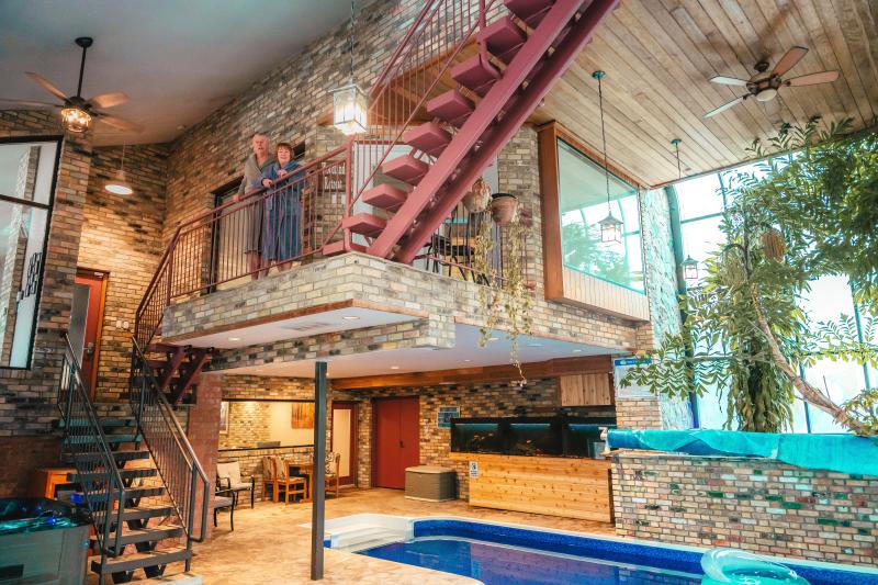 A couple wearing robes stands on an indoor balcony overlooking a pool area in a unique brick-walled retreat at Nature's Oasis Retreat. The space features a mix of rustic and modern design elements, including exposed brick walls, wooden ceilings with ceiling fans, hanging plants, and large windows letting in natural light. Below the balcony, there’s a lounge area with chairs and a dining table, and the pool is partially visible in the foreground.