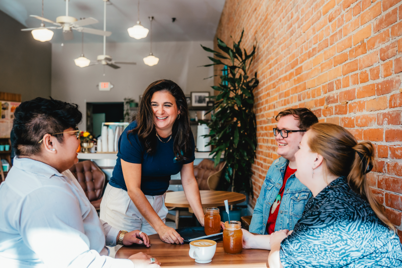 Four people sit around a table inside a cozy café with exposed brick walls and warm lighting, smiling and chatting while enjoying coffee and iced drinks. A woman standing beside the table leans in and laughs with the group, creating a welcoming and social atmosphere.