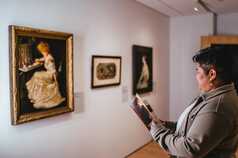 Person viewing framed paintings in an art gallery while holding a brochure, standing in front of a portrait-style artwork on a white wall.
