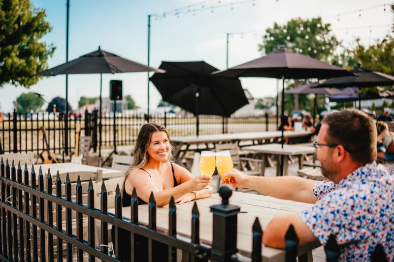 Two people smile and cheers glasses of beer while seated at an outdoor patio with umbrellas, picnic tables, and string lights in the background.